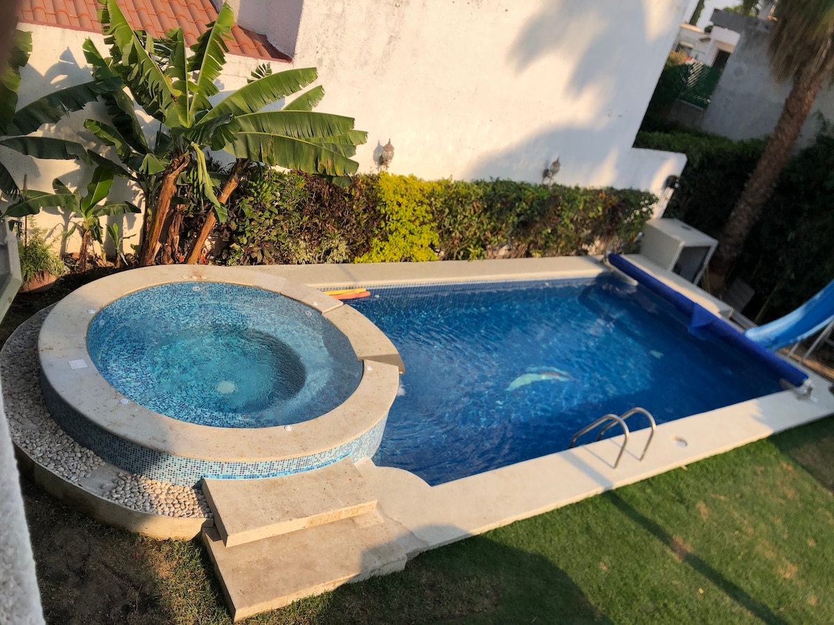 A private swimming pool is complemented by a jacuzzi, both featuring clear water. A slide integrates into the pool area, surrounded by a well-maintained garden with shrubs and palm trees providing greenery. The scene is framed by a white wall, creating a secluded atmosphere.