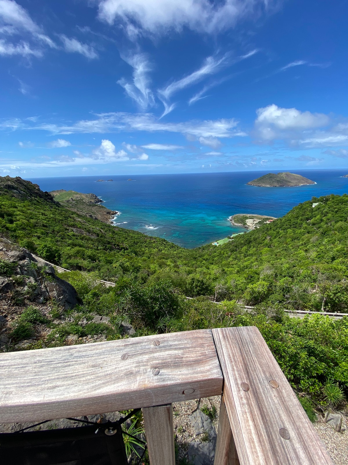 A sweeping view captures the vibrant turquoise waters of the coastline, framed by lush green hills. Islands dot the horizon under a clear blue sky, while the wooden railing in the foreground provides a picturesque viewing point of the landscape.