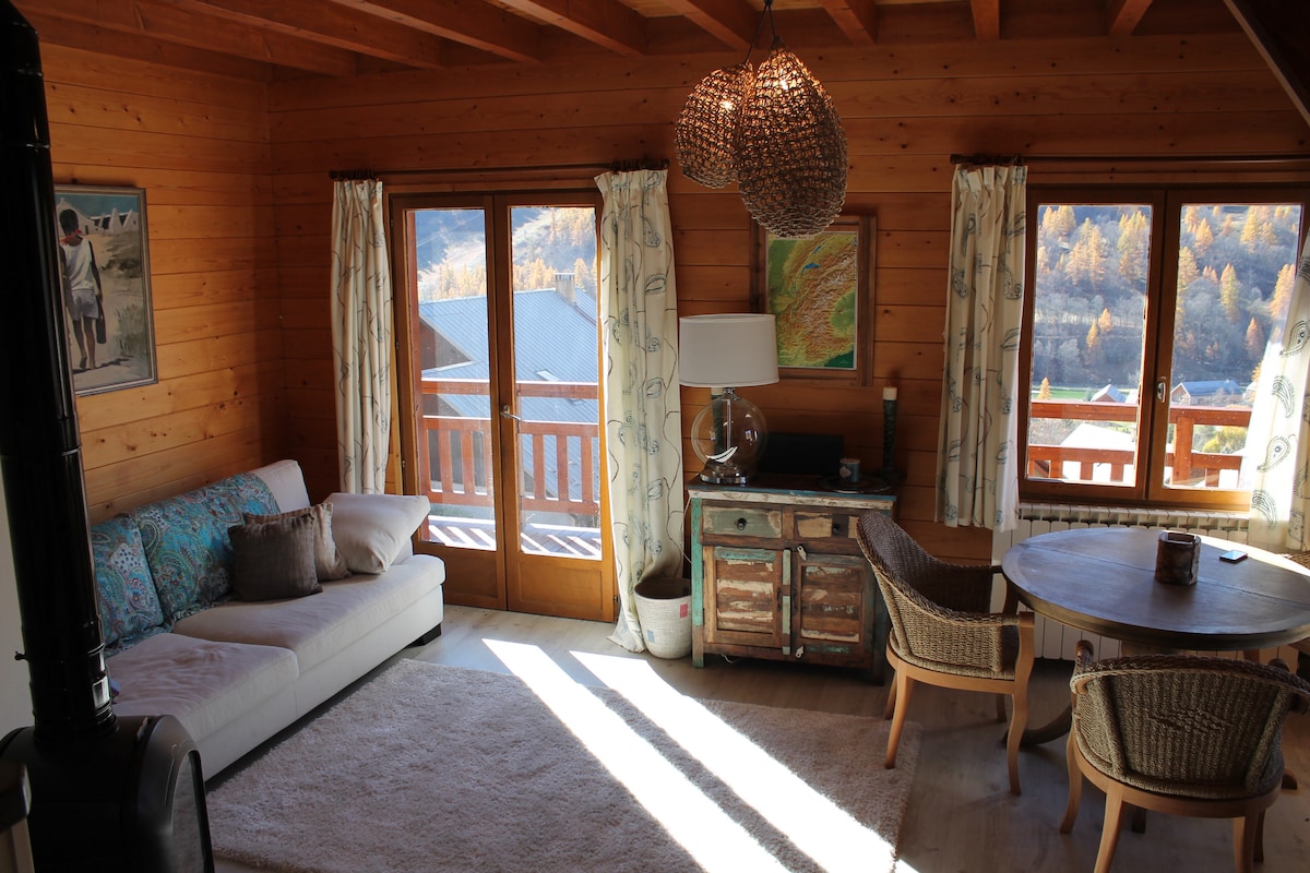 An open-plan living area features a light, cozy sofa adorned with decorative cushions, complemented by a rustic wooden dining table surrounded by woven chairs. Large windows allow sunlight to fill the space, revealing views of the village and the surrounding mountains.