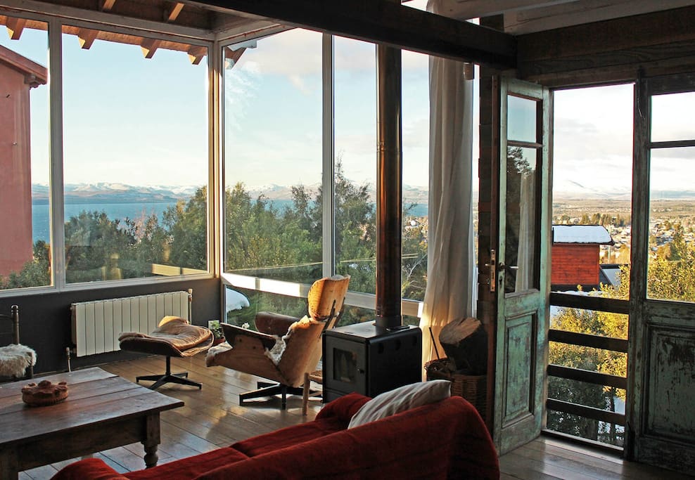 A bright living area features large windows that offer panoramic views of the landscape. Wooden flooring complements a rustic coffee table and comfortable seating. A small wood stove is positioned nearby, adding to the cozy ambiance of the space.