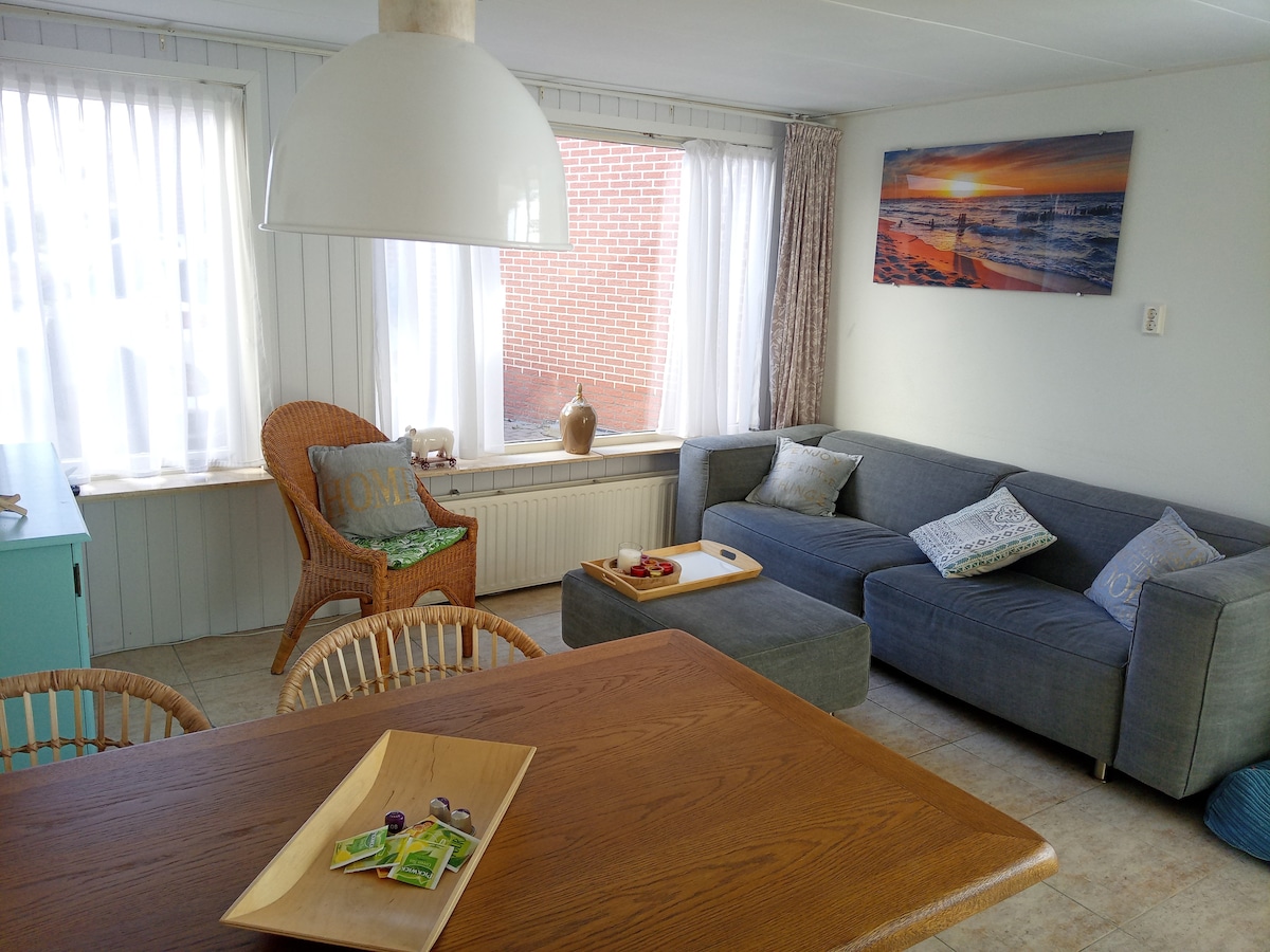 A bright living area features a cozy gray couch, a wooden dining table, and wicker chairs. Natural light filters through sheer white curtains, illuminating a decorative wall art piece above the couch. A wooden serving tray rests on the table, complemented by a selection of snacks.