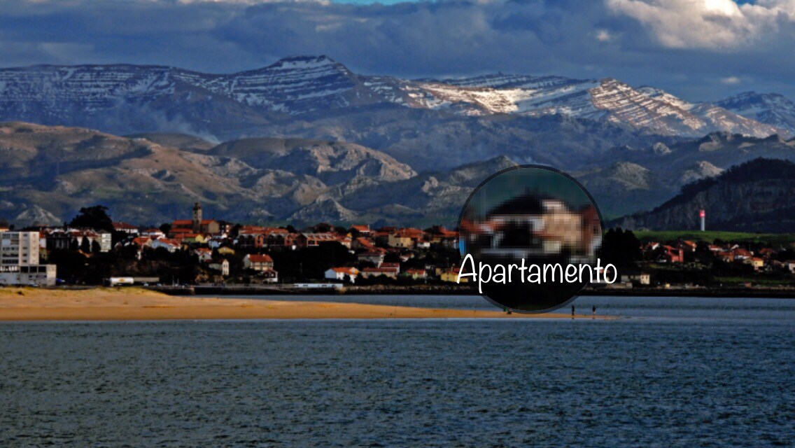 The image captures a scenic view of a coastal area with sandy shores and distant mountainous terrain, partially draped in snow. The village of Pedreña is visible in the foreground, showcasing a collection of buildings against a backdrop of natural beauty.