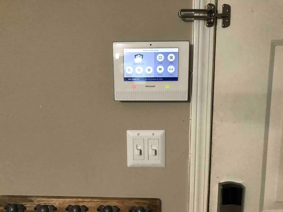 A security panel is mounted on the wall next to a light switch. The panel features a digital screen displaying icons for various functions. A set of hooks is visible below the panel, likely for hanging items such as keys or jackets.