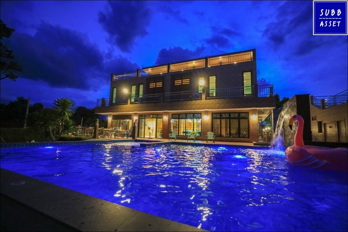 The outdoor area features a well-lit pool surrounded by a patio, showcasing turquoise water under a twilight sky. The house, with large windows, reveals an inviting interior. A pink flamingo float adds a playful touch to the serene setting.