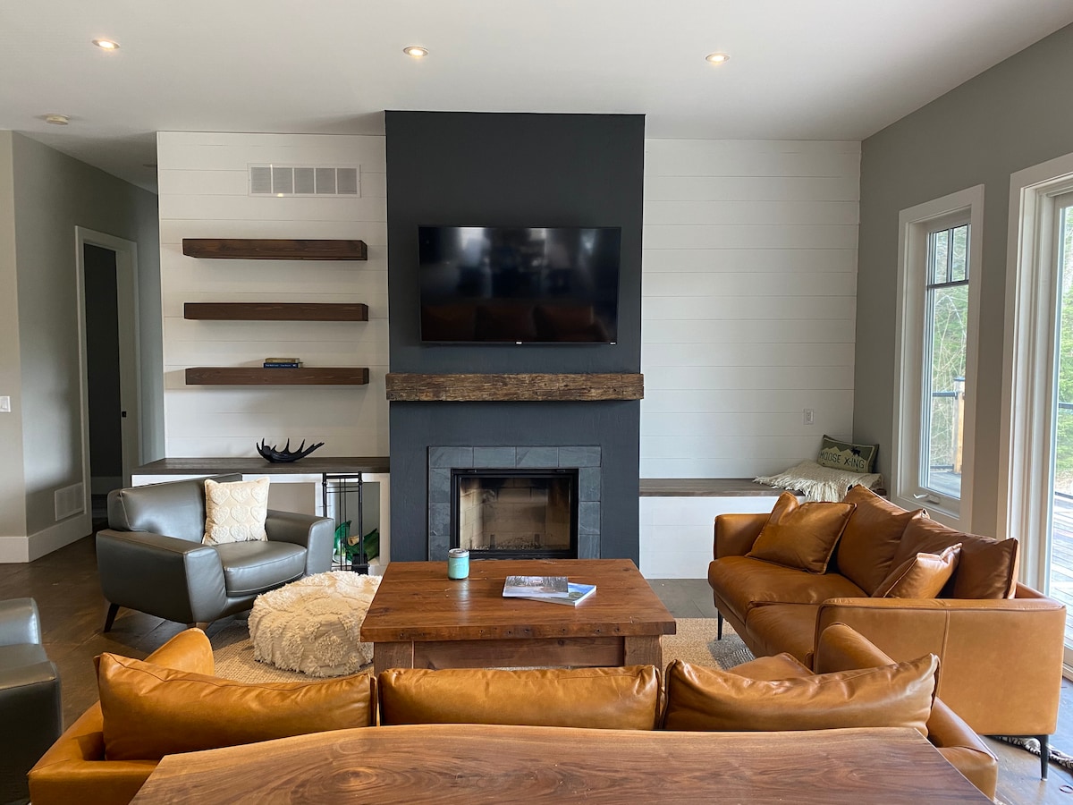 A comfortable living area features a warm color palette with a brown leather sectional sofa and a wooden coffee table. A modern fireplace is situated beneath a mounted television, and light floods in from large windows, enhancing the inviting space.