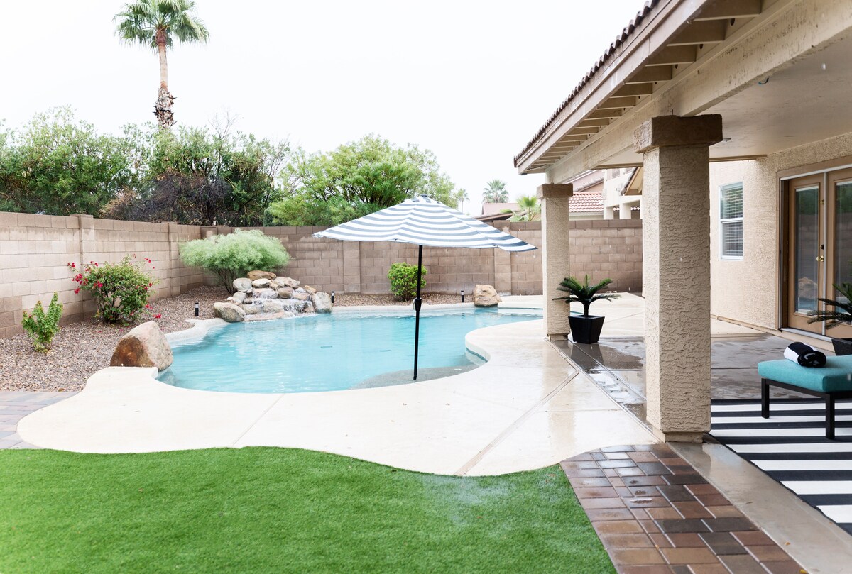 A tranquil backyard area is depicted, showcasing a curved pool surrounded by lush landscaping and a stone feature. A striped umbrella provides shade over a seating area, while a green lawn enhances the outdoor aesthetic. The patio is further adorned with neutral-colored tiles and potted plants.