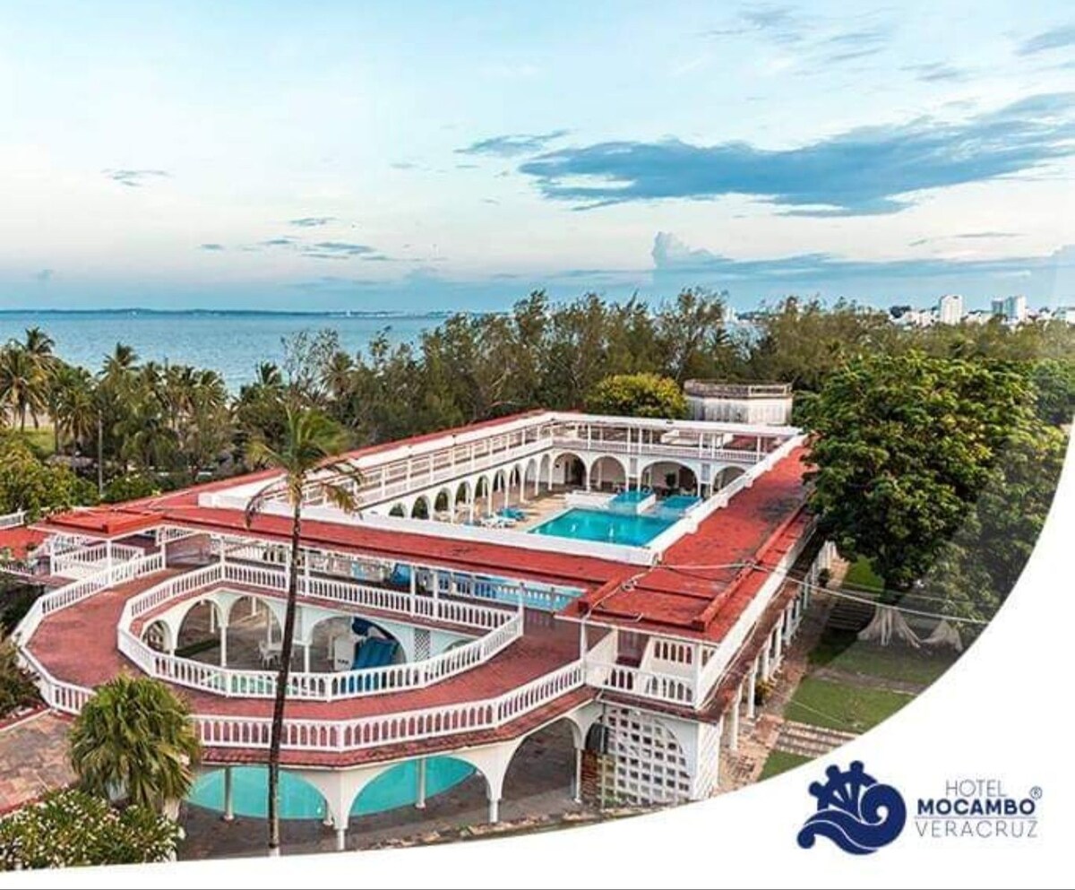 An aerial view of Hotel Mocambo reveals a spacious courtyard surrounded by trees, with two swimming pools visible. The building features classic architecture and symmetrically arranged balconies, providing an inviting atmosphere with views of the nearby ocean.