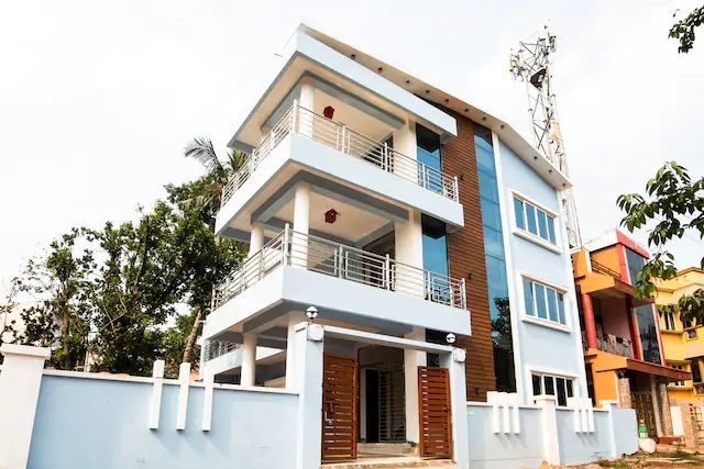The exterior of the three-story property features a modern design with a combination of light blue and brown accents. Large windows allow natural light into the interiors, while balconies provide outdoor space. Surrounding greenery enhances the overall suburban feel.