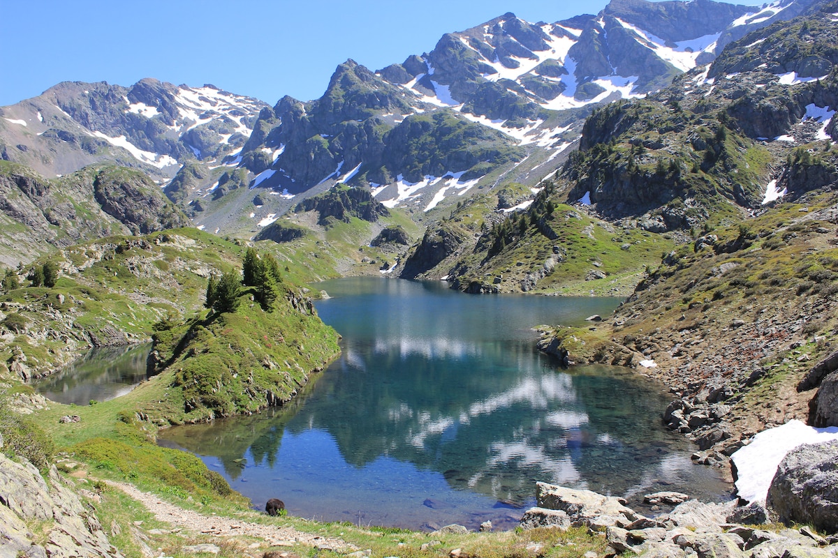 A serene alpine lake reflects the surrounding mountains and bright blue sky. Lush green hillsides circle the water, with patches of snow visible on the peaks. The peaceful landscape invites a sense of tranquility in nature.