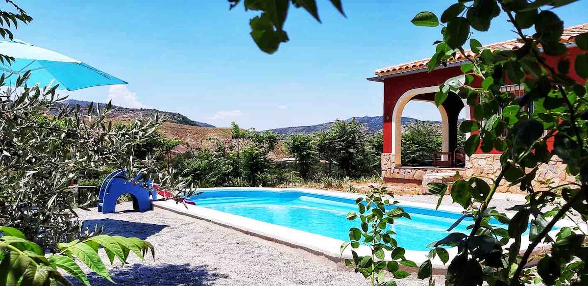 A private swimming pool is surrounded by a gravel area and views of the hills. An umbrella provides shade beside the pool, while greenery frames the view. The nearby house features a rustic facade with stone and red accents, enhancing the tranquil outdoor setting.