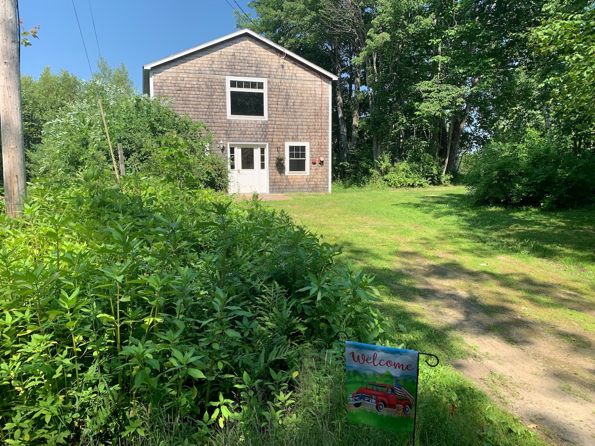 The exterior of a cottage is visible, featuring a wooden façade and multiple large windows. A 'Welcome' sign is placed on a grassy area in front, surrounded by lush greenery. The setting appears serene, with trees providing a natural backdrop.