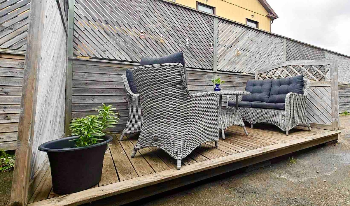 An outdoor patio is displayed, featuring a wooden deck with woven lounge furniture arranged around a small table. A decorative planter with greenery is positioned nearby, and wooden privacy panels create an enclosed feel, enhancing the cozy atmosphere of the space.