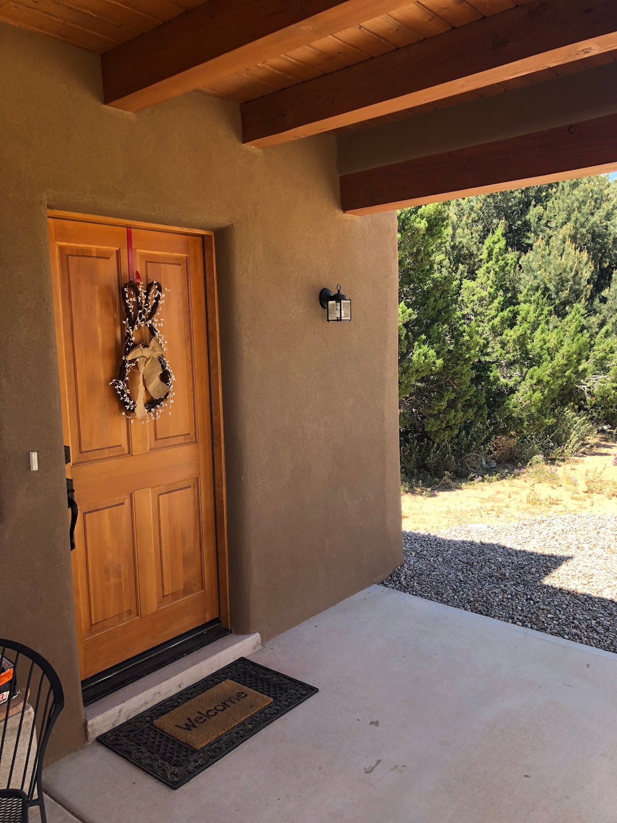 A welcoming entrance features a wooden door with a decorative wreath. A small outdoor mat with the word 'Welcome' is placed at the door, while light illuminates the entrance from a wall fixture. Natural landscaping is visible in the background.