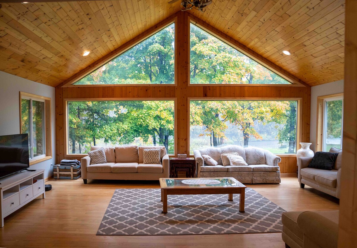 A spacious living room features large triangular windows that provide ample natural light and views of the outdoors. Two comfortable sofas are arranged around a wooden coffee table, while a soft area rug anchors the seating area. A television is positioned on a low cabinet beside the windows.