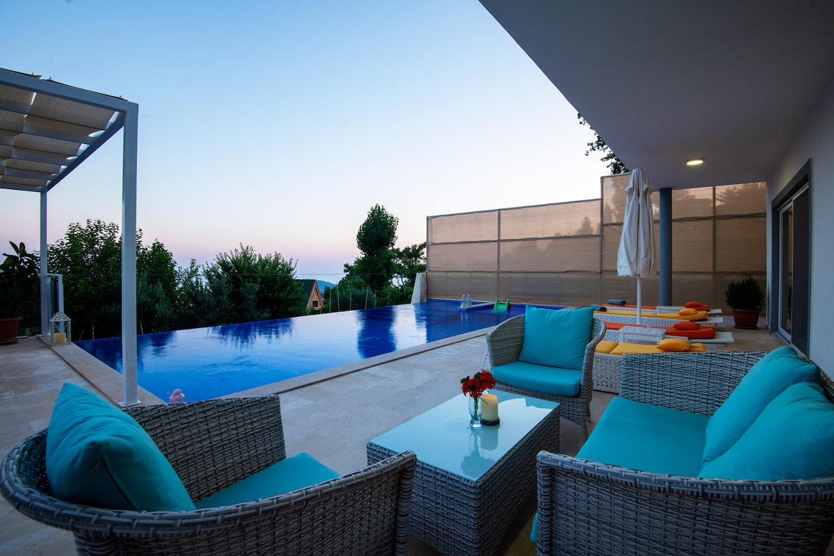 A spacious outdoor terrace is adorned with comfortable rattan seating and a glass coffee table. The shimmering infinity pool reflects the serene evening sky, bordered by vibrant greenery. Sun loungers in bright hues promise relaxation, while an umbrella provides shade for sunbathing.