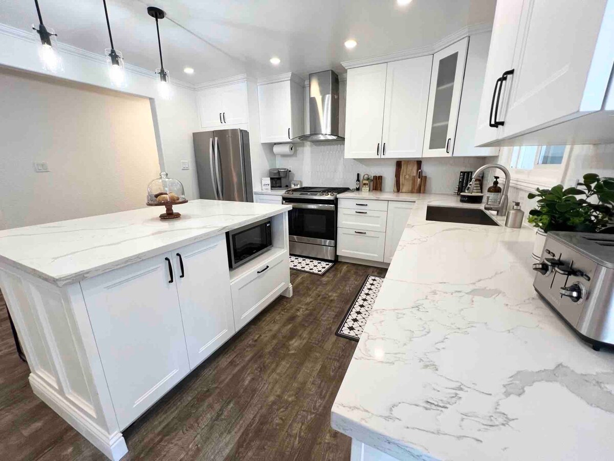 A modern kitchen is highlighted by white cabinetry and a large island featuring a marble countertop. Stainless steel appliances, including a refrigerator and a gas range, are visible. Decorative accents, such as a bowl of fruit and small potted plants, add a welcoming touch.
