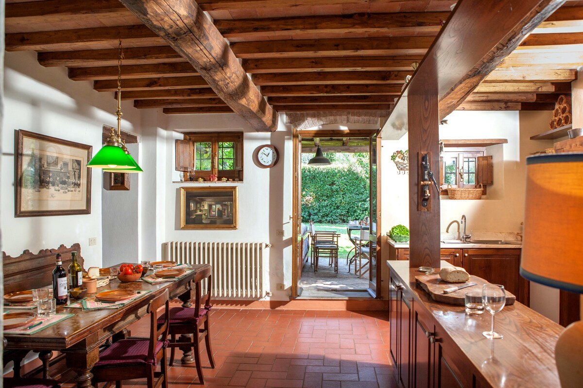 The kitchen and dining area feature rustic wooden beams and a large dining table set with plates and glassware. The space connects to an outdoor area through a doorway, while a wall clock and decorative elements enhance the warm ambiance. Glassware and a wine bottle are placed on a side counter.