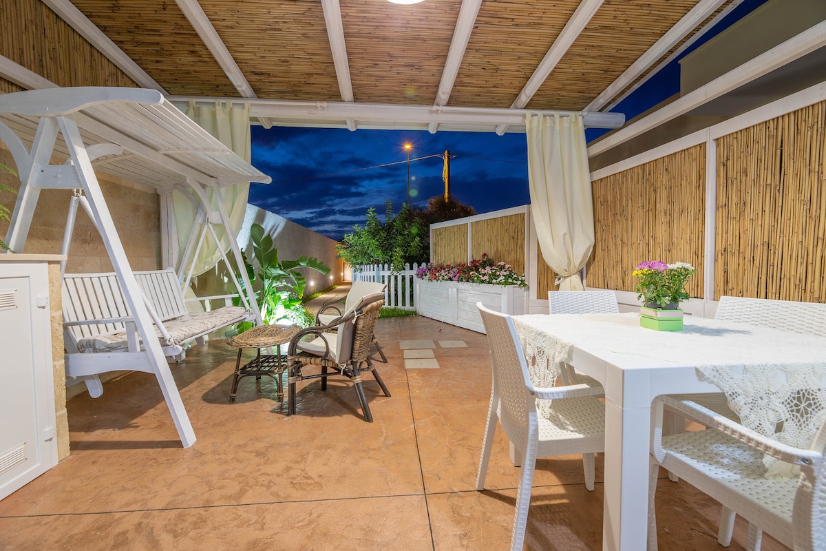 An outdoor seating area features a white table surrounded by chairs, positioned under a bamboo roof. A swing and lounge chairs are present, creating a relaxed environment. Potted plants and flowers add a touch of greenery, with lighting providing a warm ambiance as dusk approaches.