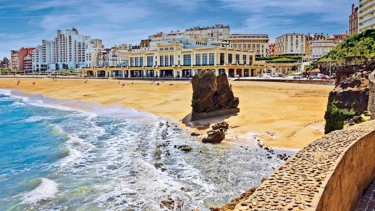 The image depicts a sandy beach with gentle waves lapping at the shore. A rock formation is visible near the waterline, with buildings lining the coastline in the background. The sky is clear with bright sunlight illuminating the scene, creating a vibrant atmosphere.