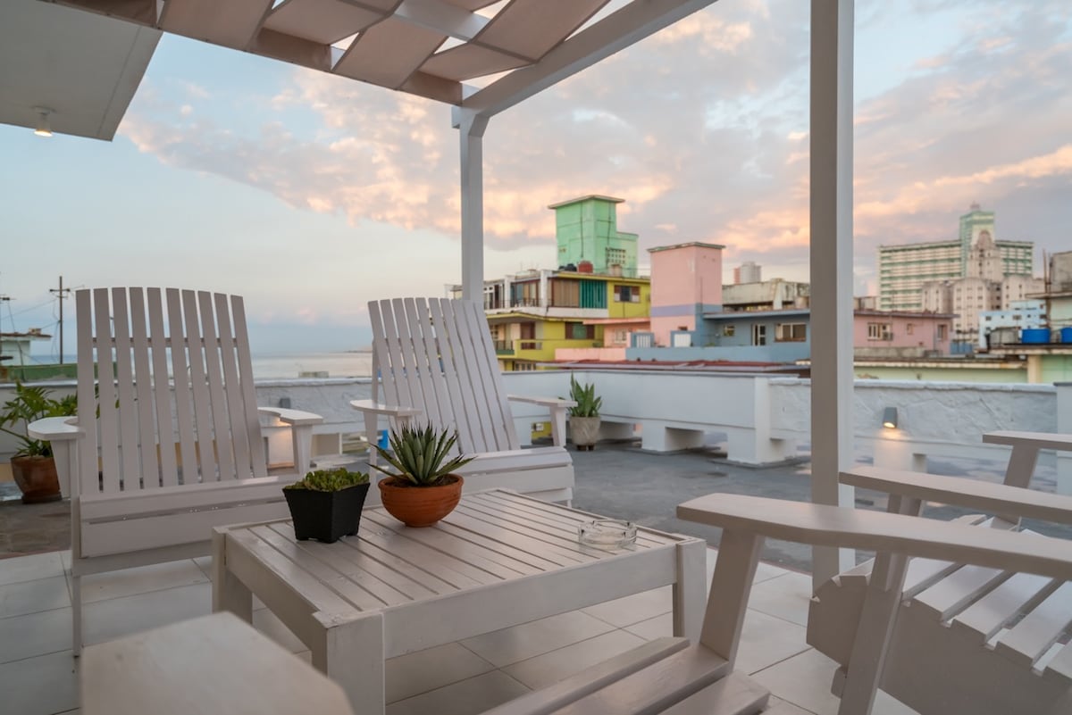 A private terrace is showcased with two white lounge chairs and a matching table. A potted plant adds a touch of greenery. The backdrop features colorful buildings and a gentle skyline, with soft hues of sunset illuminating the sky.