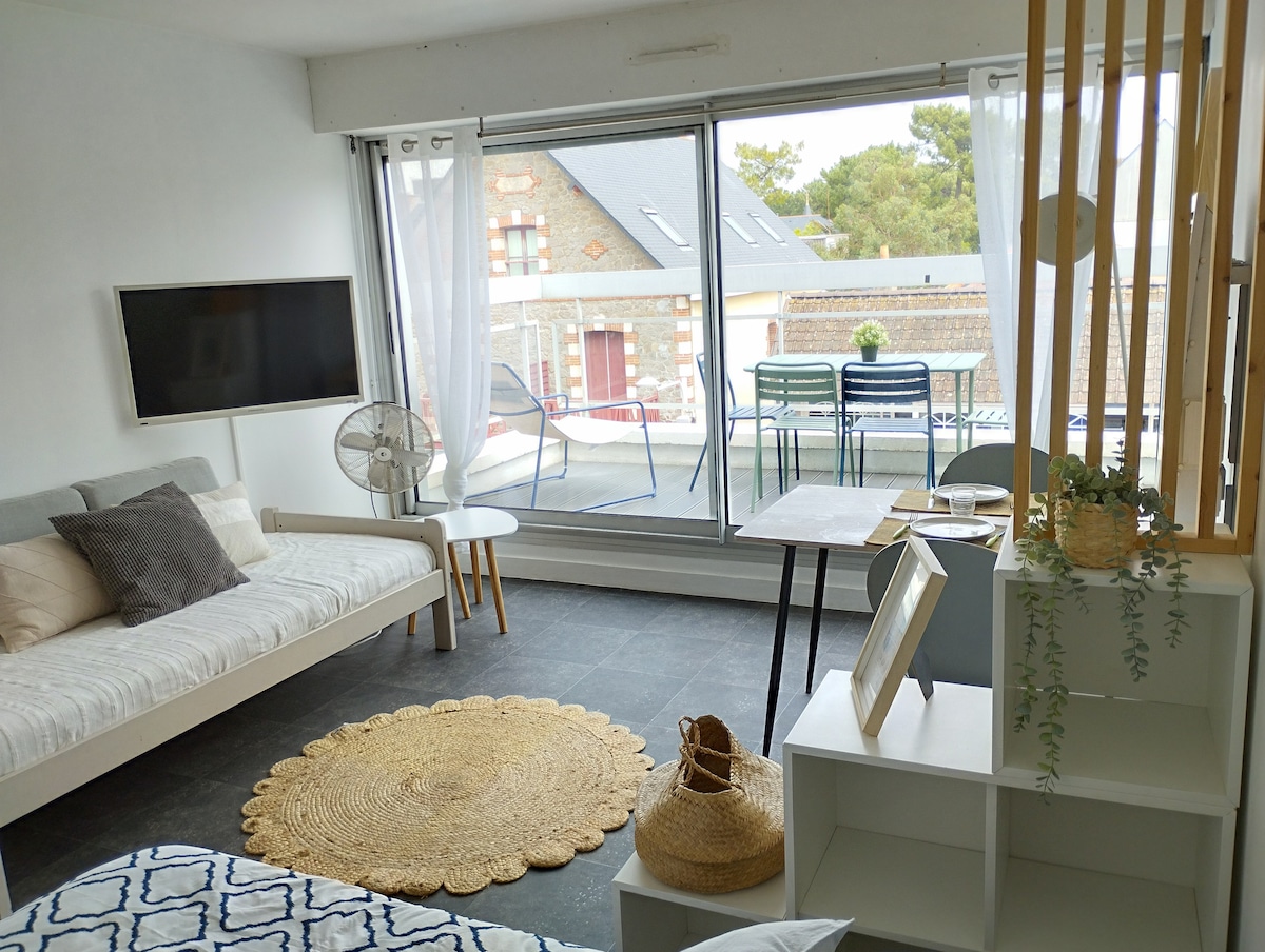 A light-filled living space features large glass doors leading to a terrace. A comfortable sofa bed is positioned next to a wall-mounted TV. A round rug lies on the floor, complemented by a small dining table and chairs. Decorative elements include indoor plants.