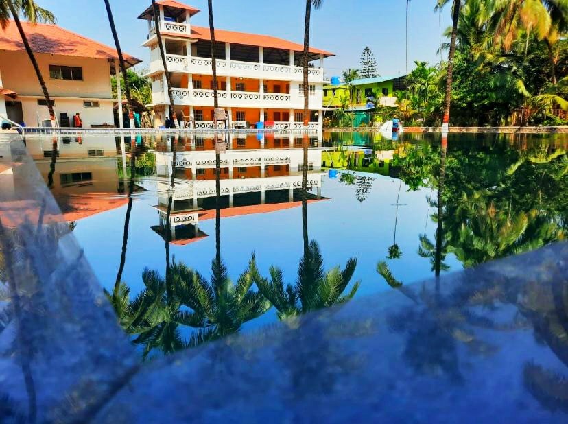 The resort is reflected in the still water of the private pool, with palm trees framing the scene. The multi-story building showcases a blend of colorful facades, while the vibrant greenery enhances the tropical ambiance. The clear blue sky complements the serene setting.