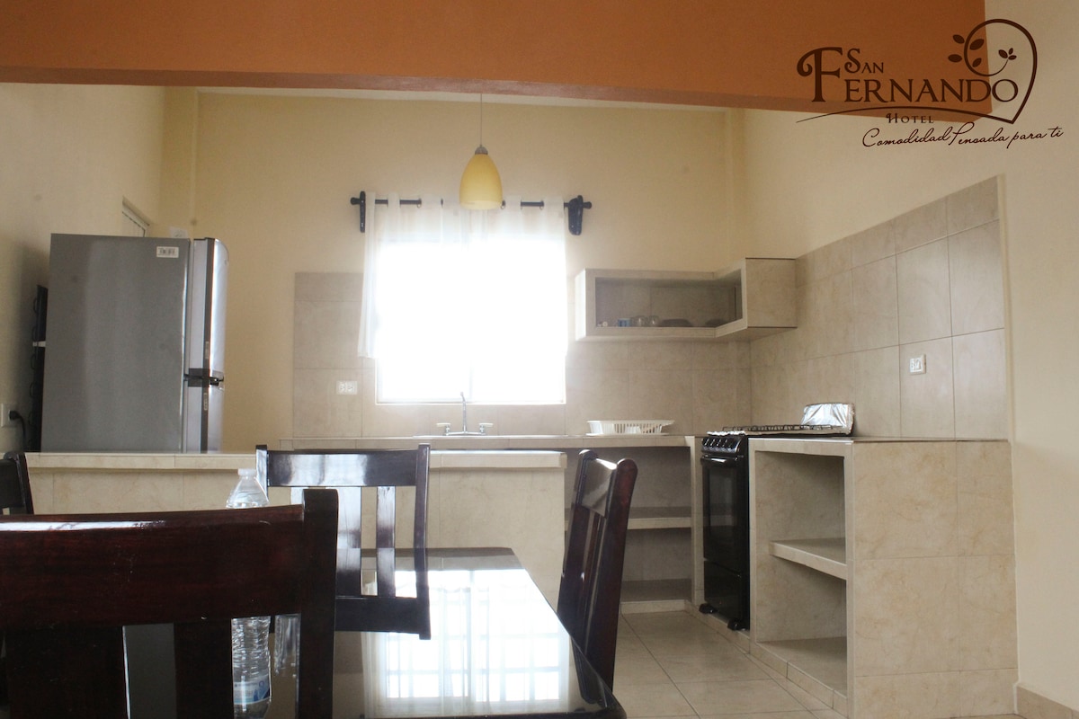 A modern kitchen is presented with neutral tile walls and light-colored cabinetry. Stainless steel appliances, including a refrigerator and stove, are visible. A minimalist dining table with dark wooden chairs is positioned in the foreground, inviting shared meals in a cozy environment.