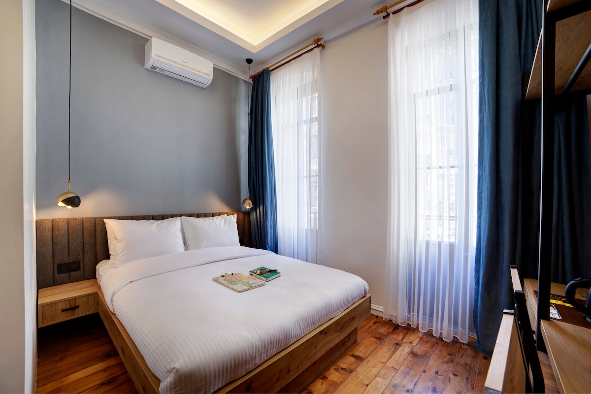 A light-filled bedroom features a spacious bed with crisp white linens, flanked by two modern bedside lamps. Soft blue curtains frame large windows, and wooden flooring adds warmth to the space. A small nightstand holds a selection of books, enhancing the overall ambiance.