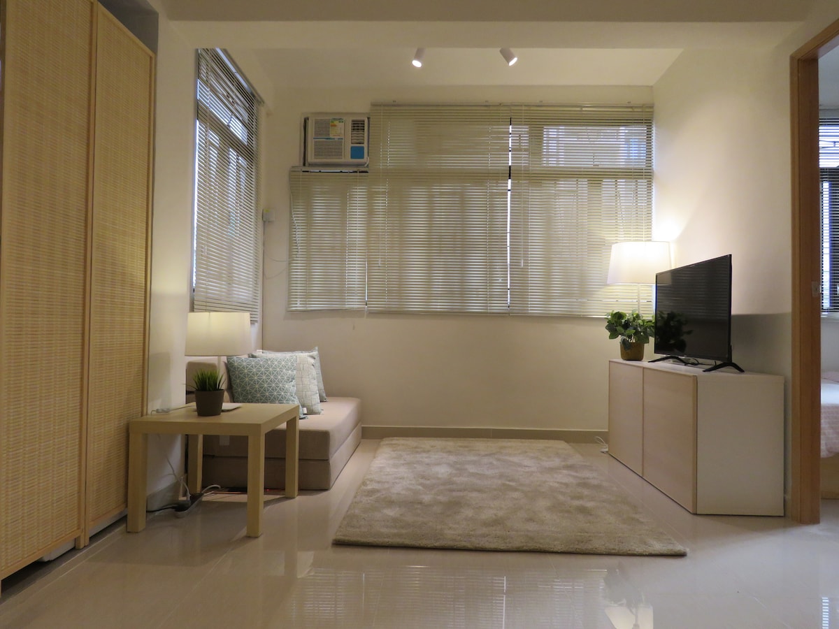 A spacious living area features a light-colored sofa and a small wooden coffee table beside a decorative plant. Natural light enters through large, partially covered windows. A contemporary TV stand holds a television, and soft lighting is provided by a nearby lamp.