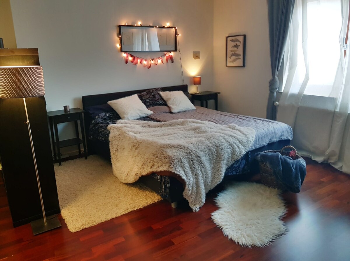The spacious bedroom features a large bed dressed in soft linens, complemented by gently glowing string lights above. A cozy rug and additional soft textiles enhance the inviting environment. Nearby, a lamp and wooden nightstands provide functional decor, while natural light filters through sheer curtains.