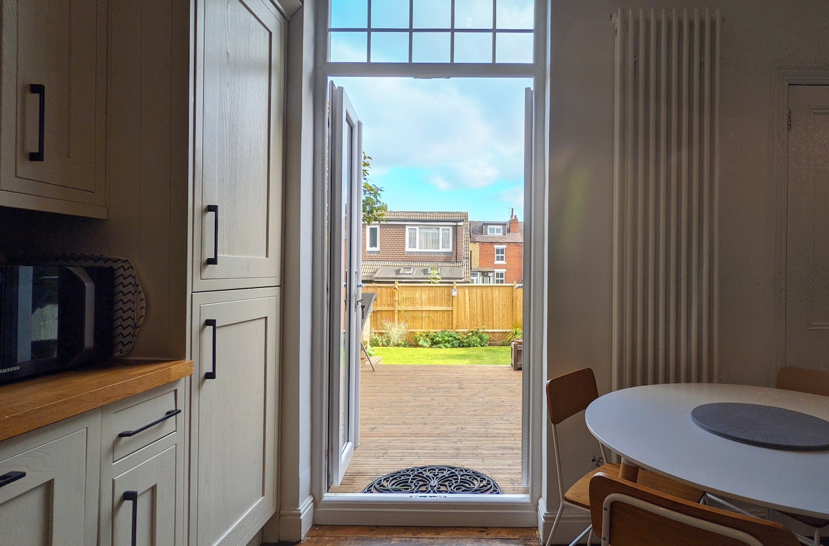 The image captures a view from the kitchen, framed by wide open French doors leading to a wooden deck. Natural light floods in through the entry, highlighting the clean lines of the cabinetry and the round dining table with wooden chairs.