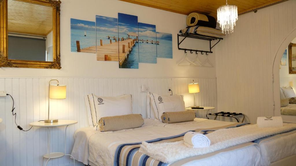 A bright room features two spacious beds with soft linens and rolled towels. Light wood accents and a large mirror enhance the space. A modern chandelier hangs from the ceiling, complemented by bedside lamps providing gentle illumination. A large artwork of a pier adds visual interest to the wall.