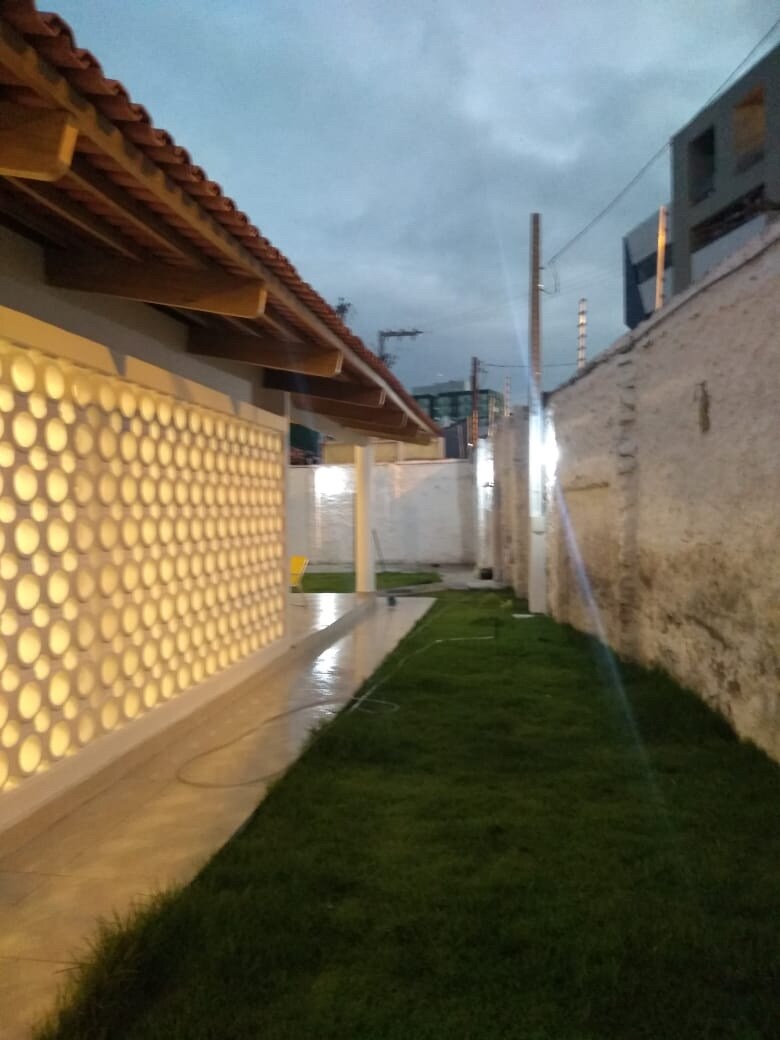 A well-maintained side yard is depicted, featuring a smooth pathway bordered by lush green grass. A wall built with cobogó blocks provides textured visual interest, while subtle lighting enhances the serene atmosphere. The space is surrounded by high walls, adding to privacy and security.