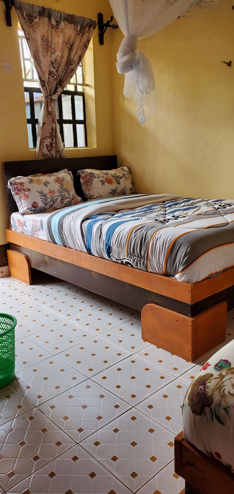 A cozy sleeping area features a wooden bed with an elegantly patterned duvet and pillows. Natural light enters through a window adorned with light curtains, illuminating the patterned tiled floor. A green wastebasket is positioned nearby, adding a pop of color.