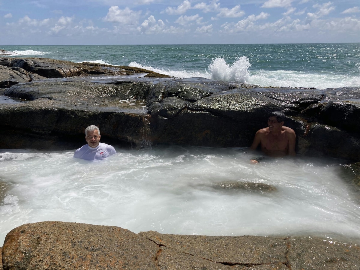 Two individuals are seated comfortably in a natural pool, surrounded by smooth rock formations. Gentle waves crash against the rocks, creating a frothy white surf. The background features a clear blue sky and the expansive view of the ocean.
