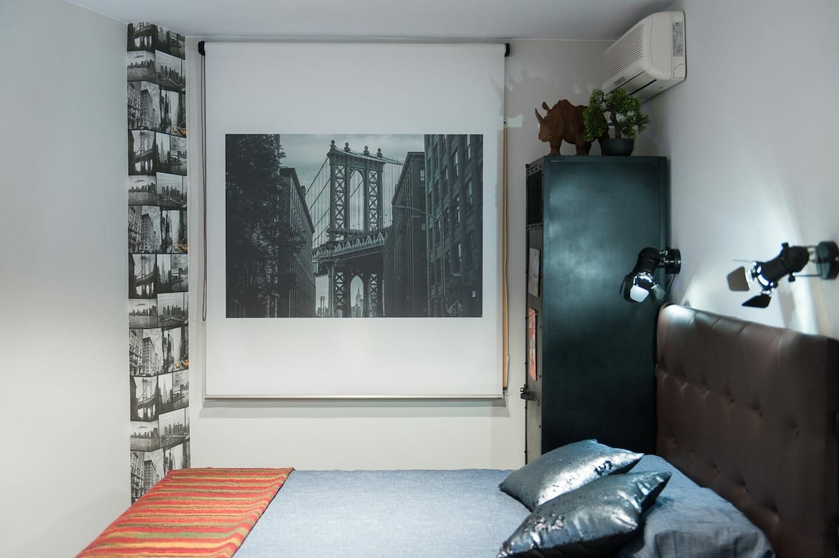 A stylish sleeping area features a prominent black-and-white photograph of a bridge, set above a dark, upholstered headboard. Decorative elements include industrial-style lighting and a subtle textured wall with cityscape imagery. A striped textile adds warmth, enhancing the overall aesthetic.