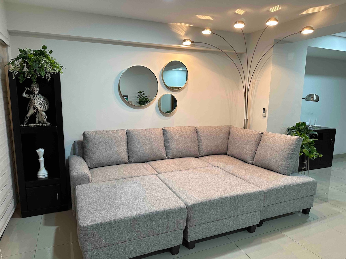 A spacious gray sectional sofa is positioned against a light-colored wall, with contemporary round mirrors reflecting ambient light above. A floor lamp provides additional illumination, while potted plants contribute greenery to the area. Decorative elements are subtly placed on either side of the sofa.