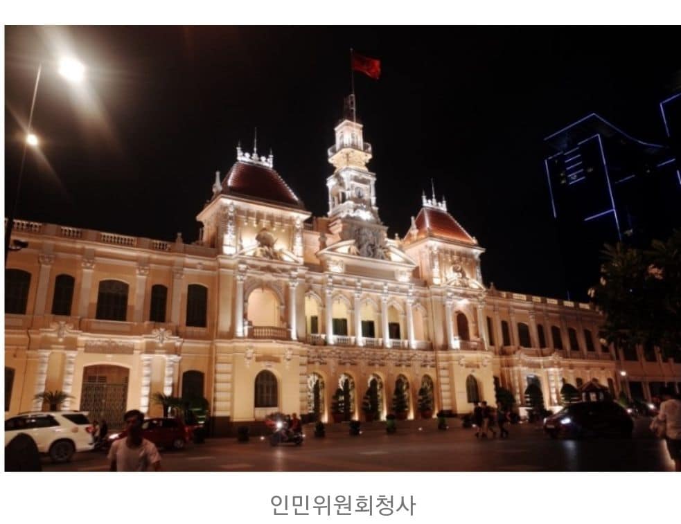 The historic People's Committee building is illuminated at night, showcasing its intricate architecture and colonial design. The structure features a tall clock tower topped with a flag and ornate details along the façade. Surrounding vehicles and pedestrians add a lively atmosphere.