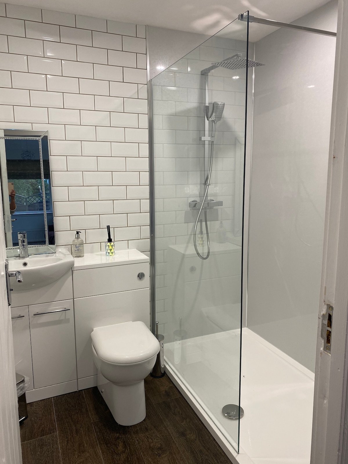A clean and modern shower room is designed with white tiled walls and a glass shower enclosure. A compact sink and a toilet are visible, with a small mirror reflecting light. Dark wood flooring adds contrast to the bright atmosphere.