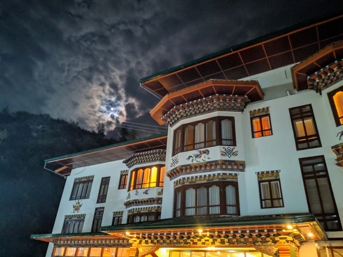 The hotel exterior is illuminated under a night sky, revealing intricate architectural details. Large windows reflect the moonlight, while the roof features traditional designs. Surrounding hills are faintly visible against the cloudy backdrop, adding to the serene atmosphere.