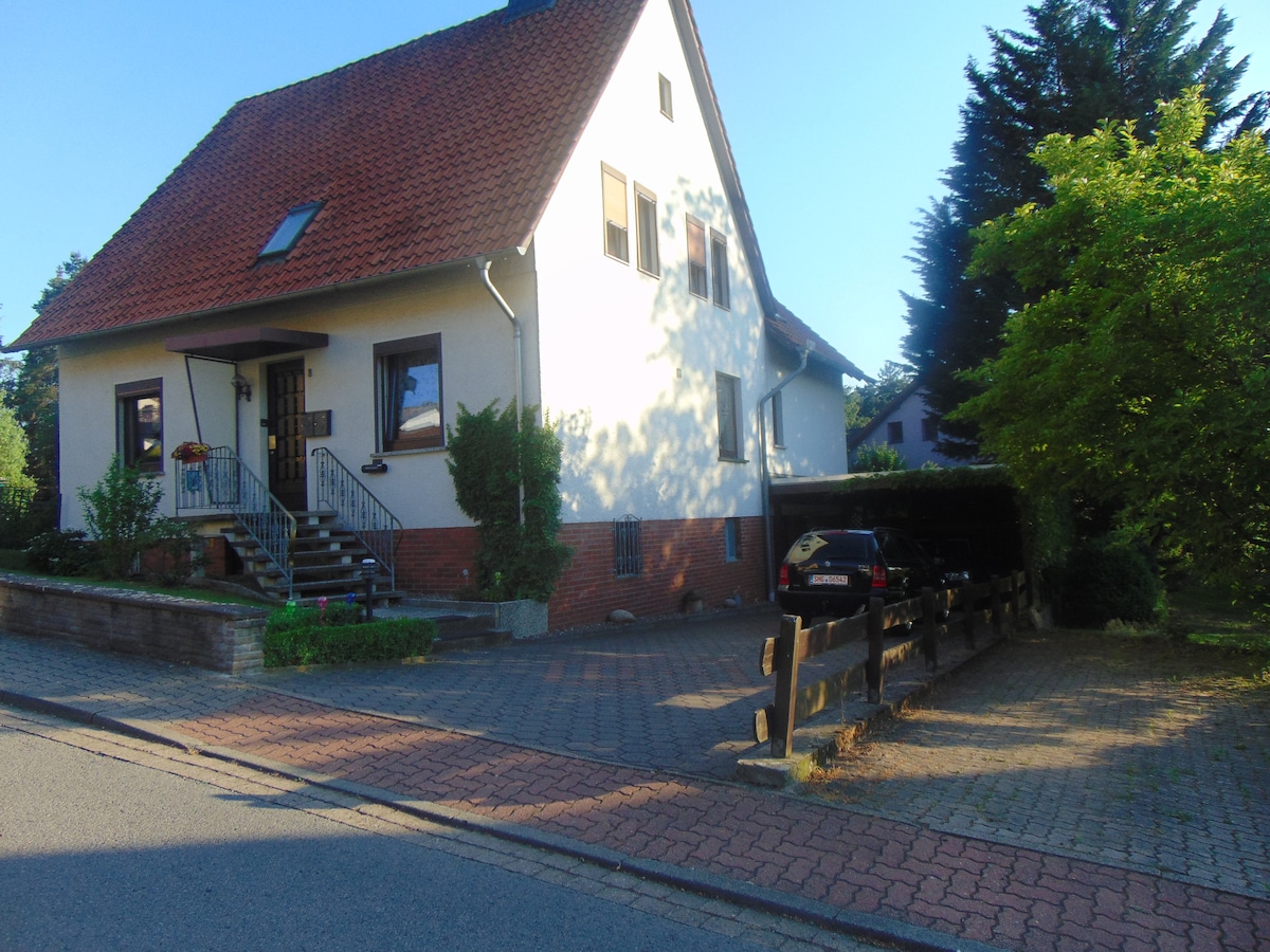 A two-story house is shown, featuring a traditional peaked roof and white exterior walls. A set of stairs leads to the front entrance, surrounded by green foliage. A brick driveway accommodates parked vehicles, while a wooden fence borders the property, set against a clear blue sky.