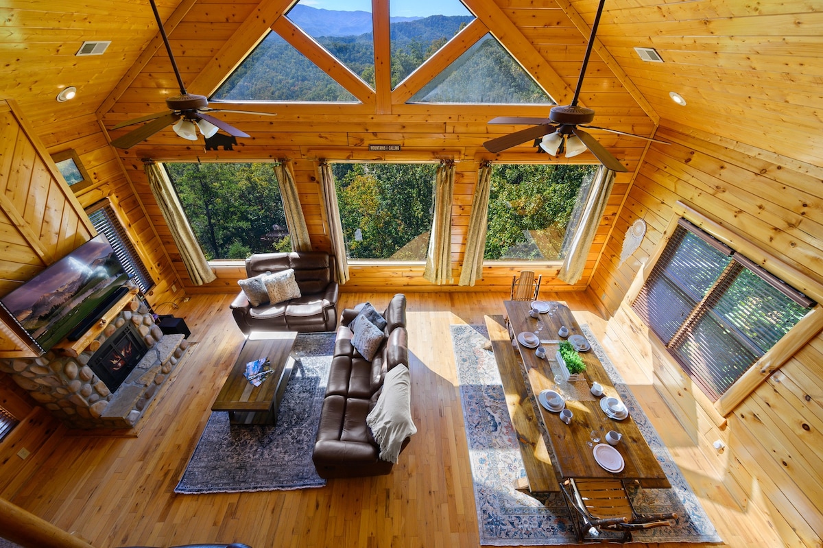 A spacious living area features large windows that provide picturesque views of the mountains. Natural wood elements dominate the space, with a comfortable sofa and a rustic coffee table. A dining table set for meals sits nearby, while a stone fireplace adds a cozy focal point.