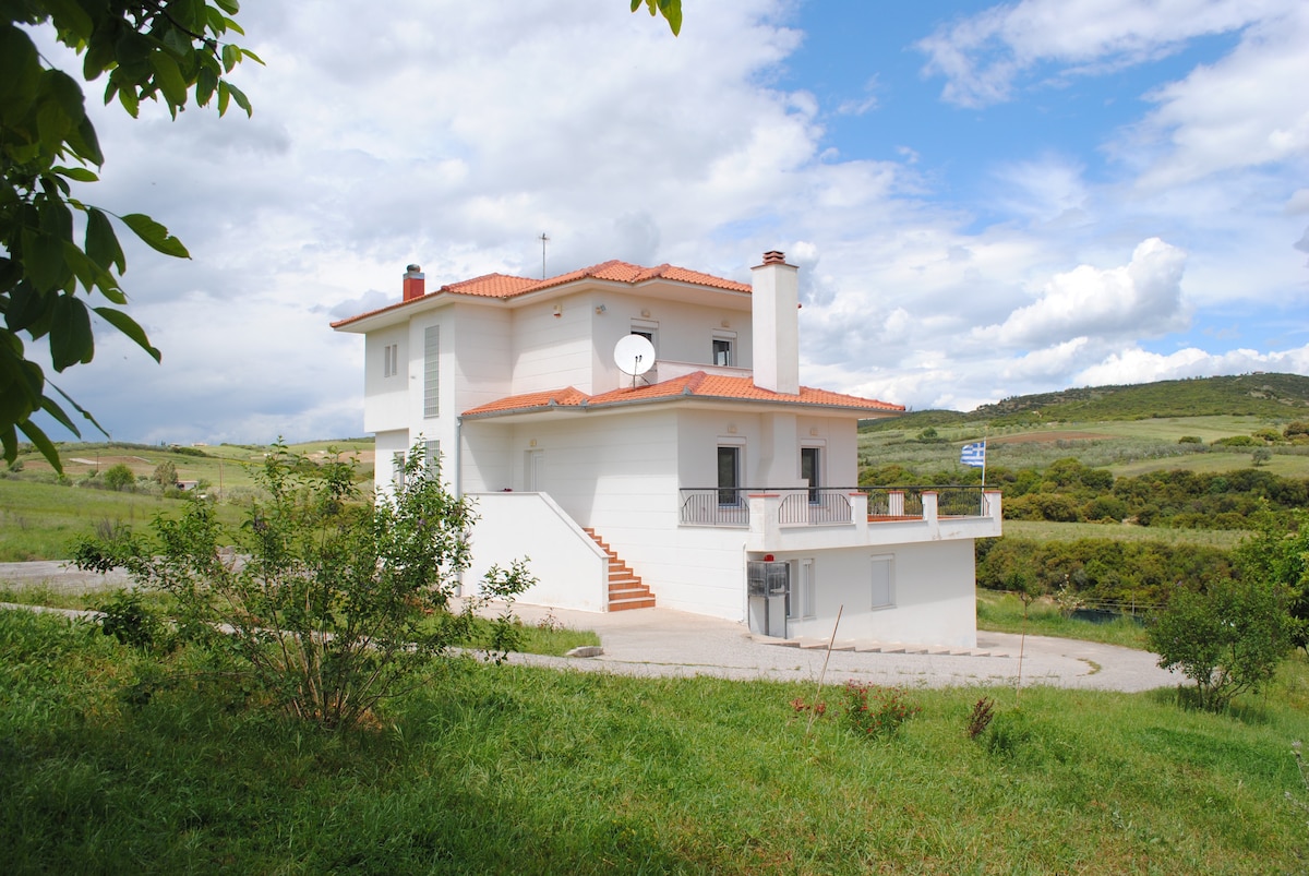 A modern three-story villa is depicted, featuring a white exterior and orange roof tiles. Staircases lead to the upper levels, and a satellite dish is mounted on the side. Lush greenery surrounds the property, complementing the serene landscape in the background.