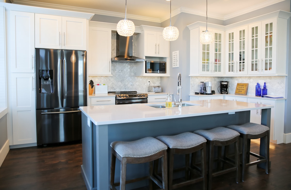 A modern kitchen is featured with sleek white cabinets and coordinating granite countertops. Stainless steel appliances, including a refrigerator and range, are present. A central island with seating for four is highlighted, beneath stylish pendant lighting. Glass-front cabinets display decorative items.