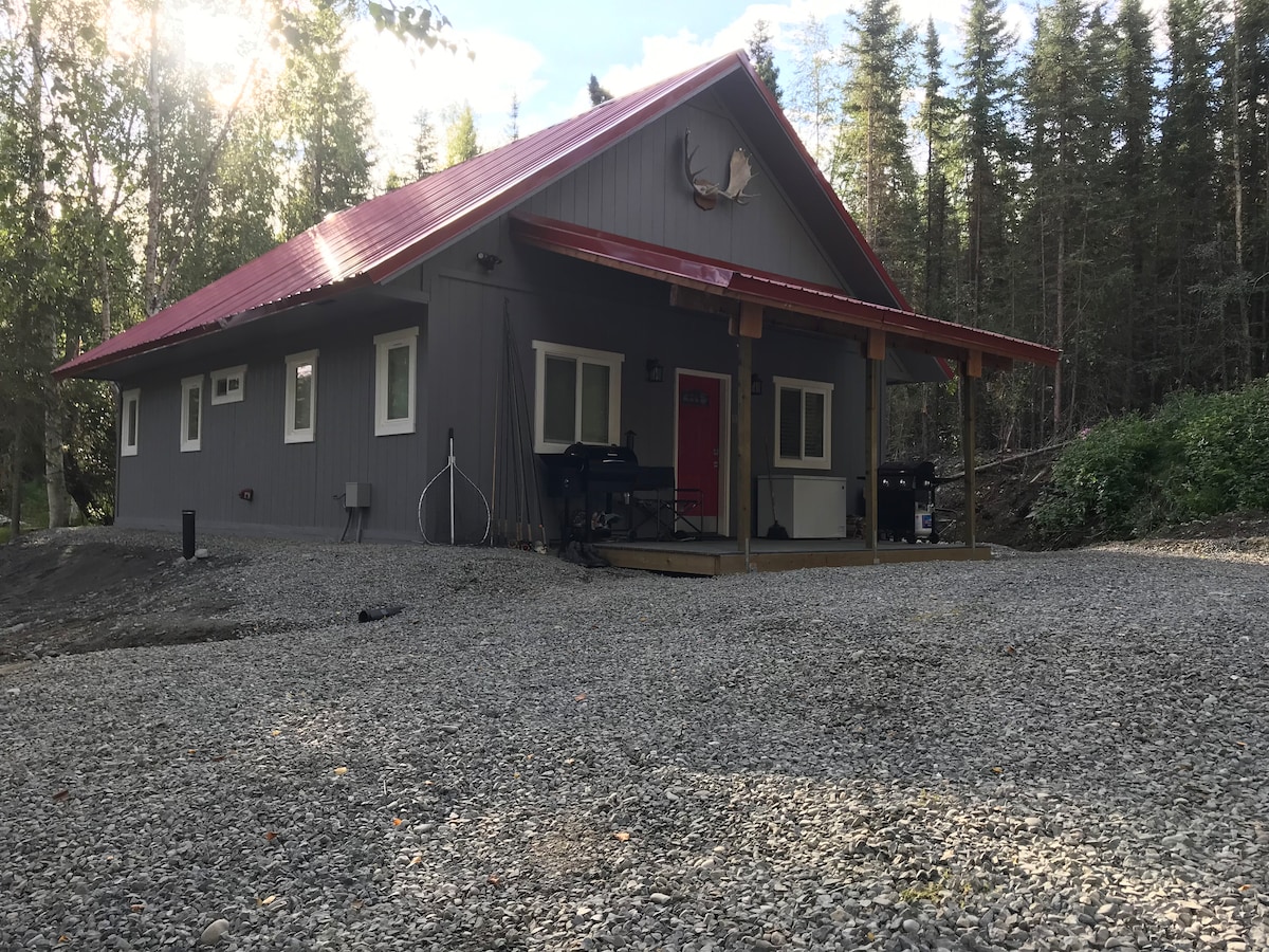 A newly constructed cabin is set amidst a wooded area, featuring a red metal roof and a front porch. Large windows provide ample natural light, while gravel surrounds the foundation. The entrance is marked by a red door, and outdoor grills are positioned nearby.