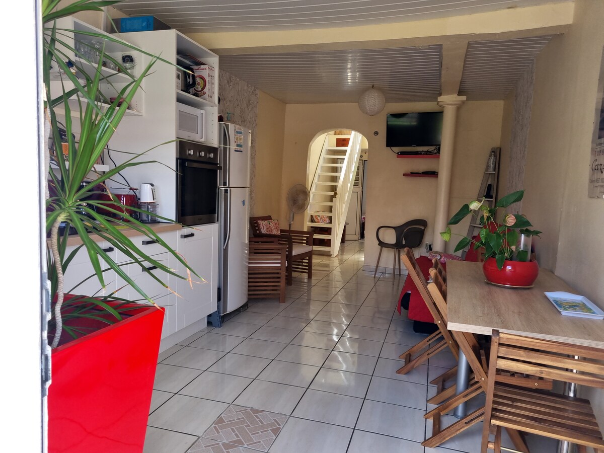 A bright dining area features a wooden table surrounded by folding chairs, with decorative plants adding a touch of greenery. A kitchenette, equipped with modern appliances, is visible on the left. Light tiles enhance the space, leading to a staircase in the background.