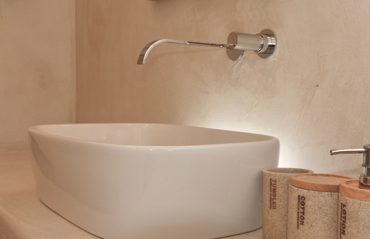 A modern bathroom is highlighted by a stylish, white basin positioned above a smooth, neutral-toned surface. A chrome faucet extends from the wall, and subtle lighting enhances the wall's texture. Nearby, organic cotton products are presented in natural-toned containers.