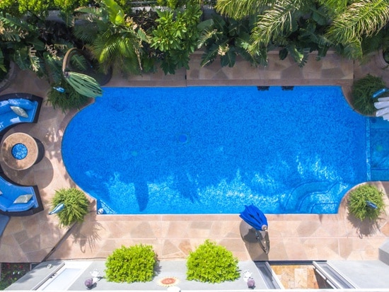 An aerial view of a lushly landscaped pool area is presented, featuring a sparkling blue pool surrounded by tropical vegetation. Comfortable seating and a circular table are visible nearby, inviting relaxation in a serene outdoor setting.