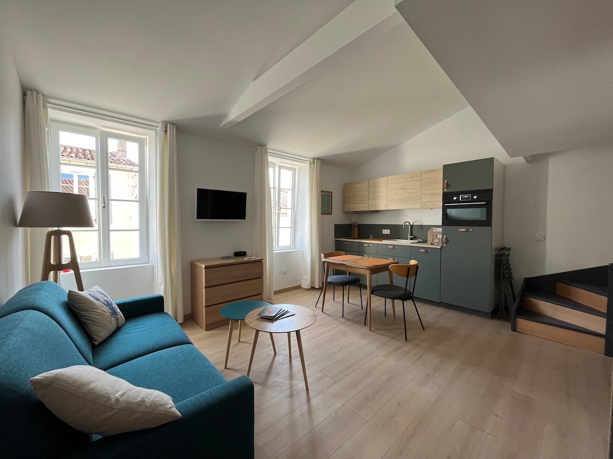 A modern studio space features a cozy blue sofa and a wooden coffee table. Natural light floods through large windows, illuminating the open kitchen area equipped with essential appliances. A stylish dining table with four chairs is positioned nearby, creating a functional living space.