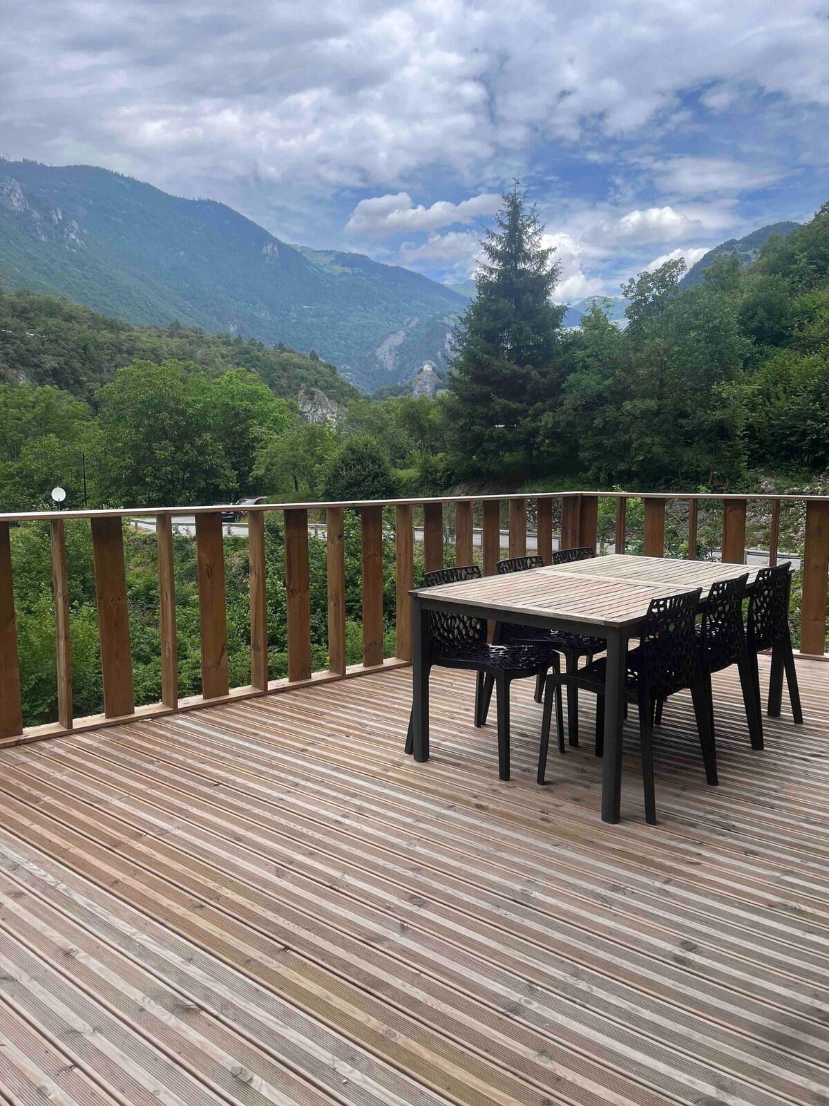 A wooden terrace features a dining table surrounded by four chairs, with scenic mountain views in the background. Lush greenery is visible below, showcasing the peaceful natural surroundings.